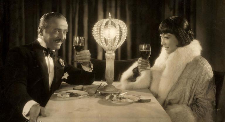 A promotional still for Pavement Butterfly showing Gaston Jacquet and Anna May Wong, seated at a dining table, raising their glasses to toast.